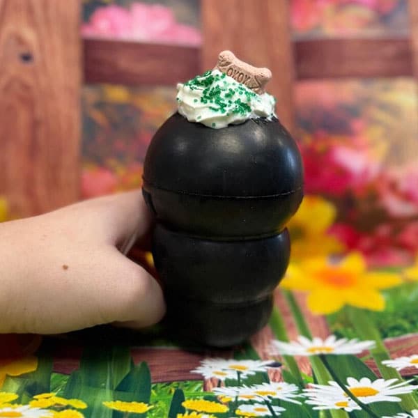 A hand holds a black dog toy filled with cream, topped with green sprinkles and a small dog bone, set against a colorful, flower-decorated background.